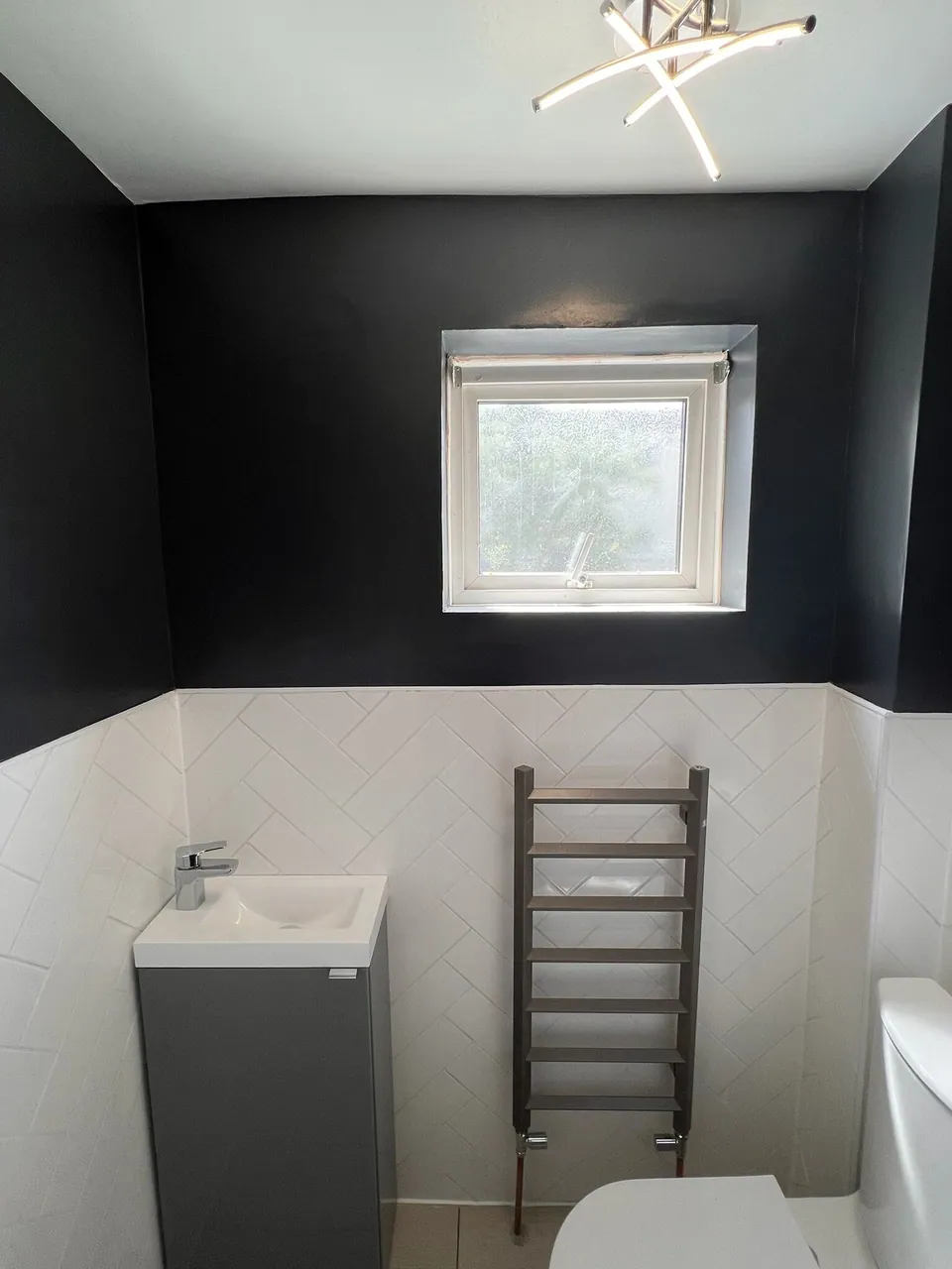 Finished cloakroom with white herringbone tiles to half height, navy painted walls, compact grey vanity and chrome towel rail, fitted by Basingstoke Bathrooms