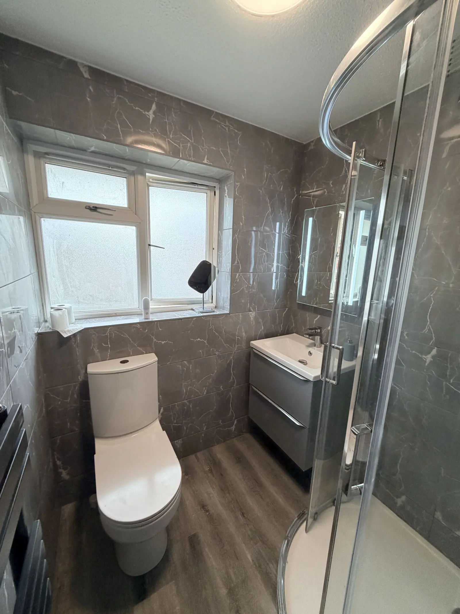 Bathroom in grey marble-effect tiles with a quadrant shower enclosure, grey vanity and black towel rail, fitted by Basingstoke Bathrooms
