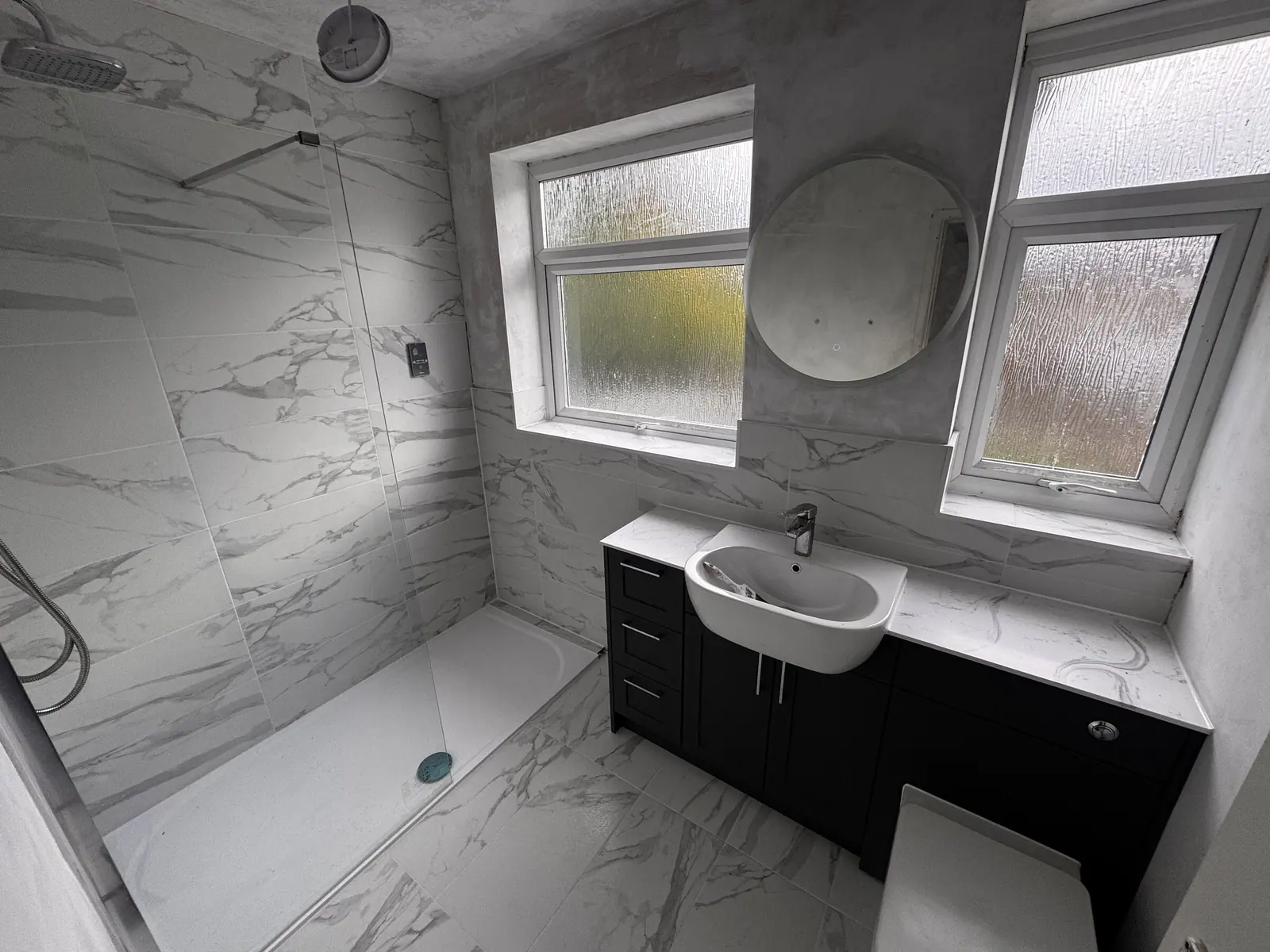 Shower room tiled in white marble-effect with a walk-in shower, navy vanity and round mirror, fitted by Basingstoke Bathrooms